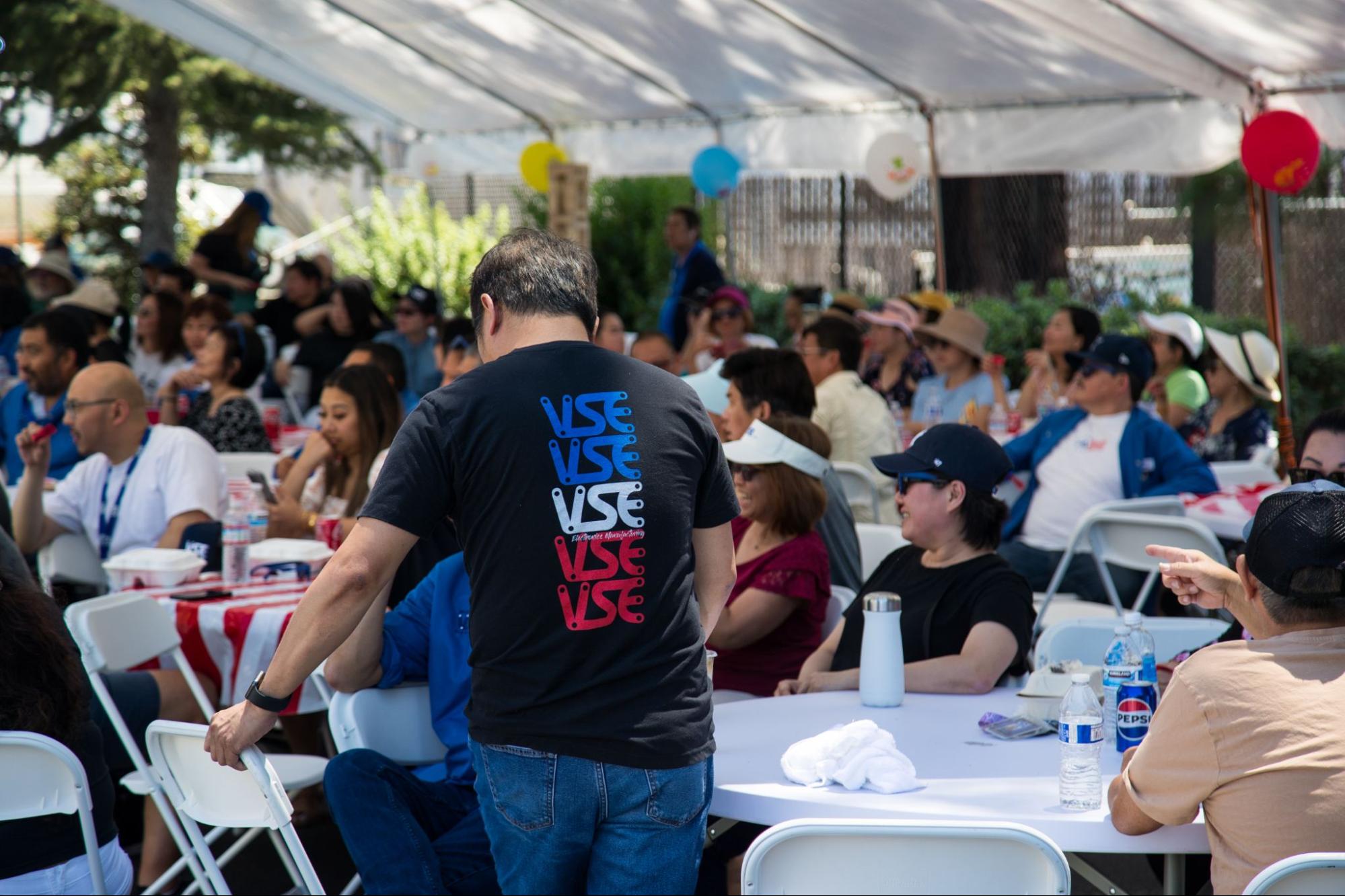 A man in a black t-shirt with a red, white, and blue "VSE" logo stands at an outdoor event.